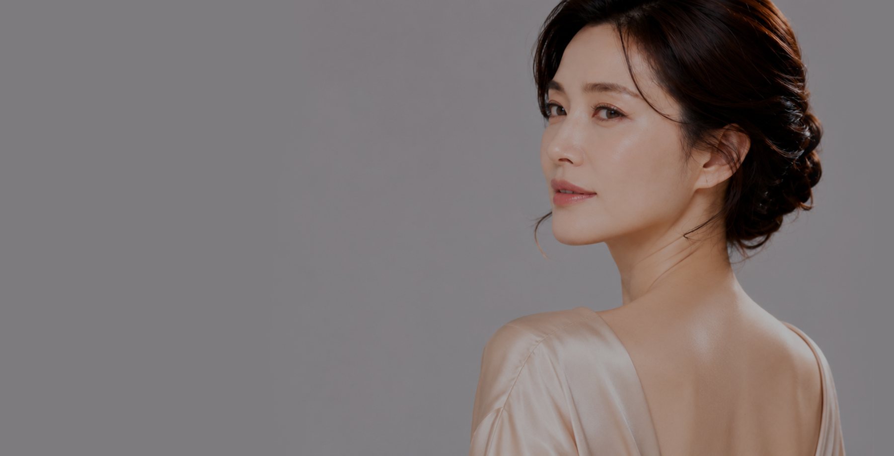 회색 바탕, 등이 파인 샴페인색 새틴 드레스로 뒤돌아보는 올림머리 중년 여성 모델